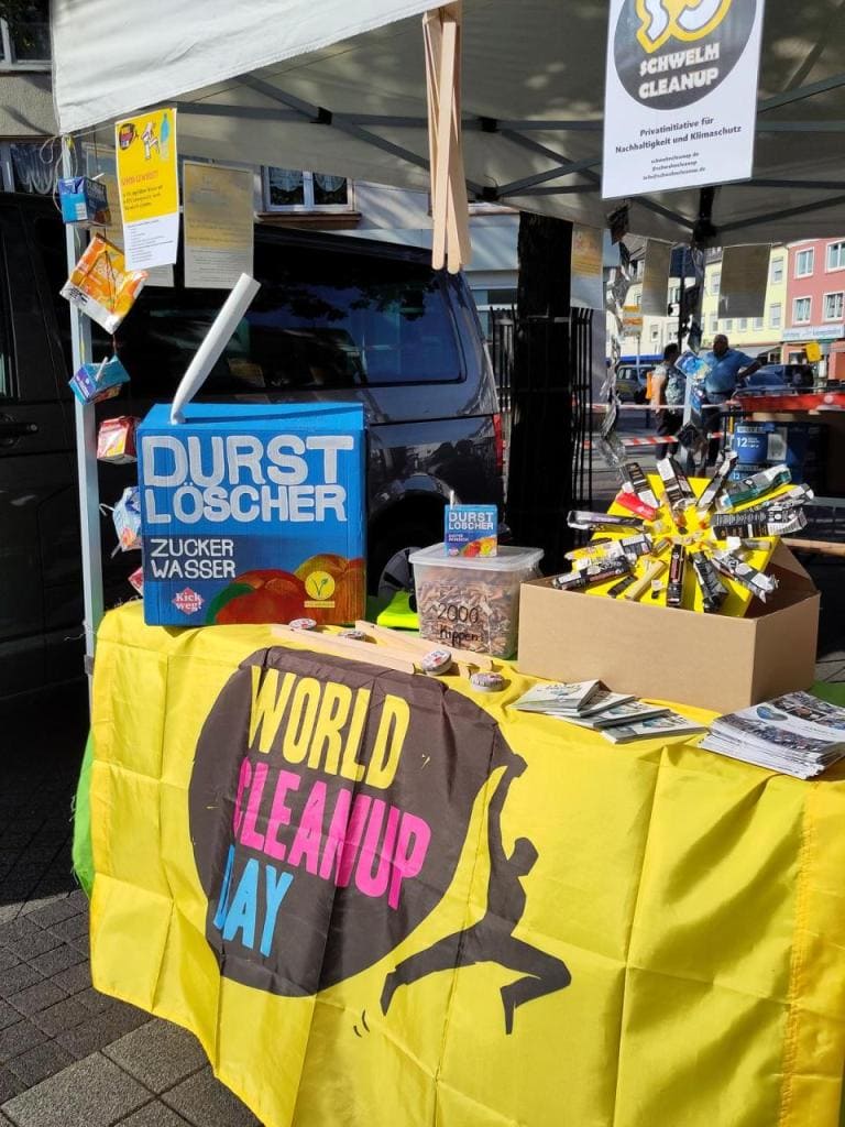 Infostand mit gelber World-Cleanup-Day-Tischdecke, einem Schild „Durstlöscher – Zuckerwasser“ und Infomaterial