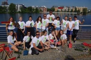Eine Gruppe in weißen Shirts mit grünem Logo posiert mit Besen, Schaufeln und Rechen auf einer Uferpromenade an der Spree.