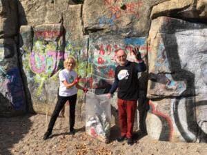 Eine Frau und ein Mann in World-Cleanup-Day-Shirts halten einen vollen Sack vor einem bunt besprühten Felsen und winken.