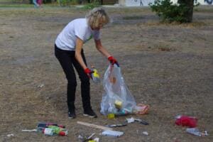 Eine Frau mit roten Handschuhen sammelt Dosen und Verpackungen von einer trockenen Wiese, um sie herum liegt noch mehr Müll.