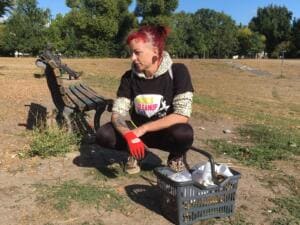 Eine Frau mit roten Haaren im World-Cleanup-Day-Shirt hockt mit roten Handschuhen neben einem Einkaufskorb voller Müll an einer Parkbank.