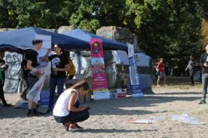 Vor blauen Pavillons und Roll-ups an Graffiti-Felsen holen Helfer Säcke ab, eine Frau hockt vorn und schaut auf ihr Handy.