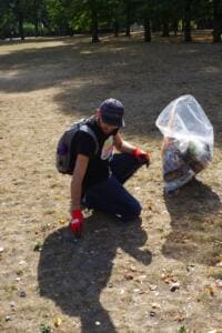 Ein Mann mit Kappe und roten Handschuhen kniet auf dem Boden und sammelt Kronkorken auf, neben ihm ein voller Sack.