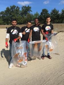 Vier junge Männer in World-Cleanup-Day-Shirts stehen mit vollen durchsichtigen Säcken auf einer Sandfläche im Park.