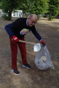 Ein älterer Mann im World-Cleanup-Day-Shirt hebt mit einem Greifer einen Plastikbecher auf und hält einen vollen Sack.