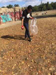 Ein Mann im World-Cleanup-Day-Shirt sammelt mit einem durchsichtigen Sack Müll auf einer laubbedeckten Wiese vor Graffiti-Felsen.