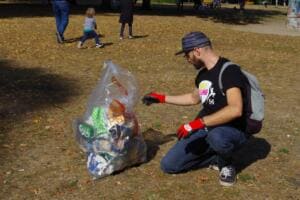 Ein Mann im World-Cleanup-Day-Shirt hockt neben einem prall gefüllten durchsichtigen Sack auf einer laubbedeckten Wiese.