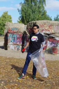 Ein Mann im World-Cleanup-Day-Shirt geht mit einem durchsichtigen Sack über eine laubbedeckte Fläche vor Graffiti-Felsen.