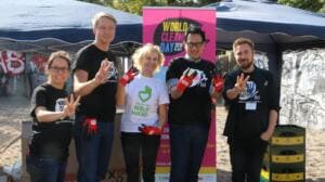 Fünf Erwachsene in World-Cleanup-Day-Shirts stehen vor einem pinken World-Cleanup-Day-Roll-up und zeigen ihre roten Handschuhe.