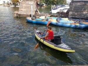 Eine Frau in Schwimmweste paddelt in einem Schlauchkajak mit einem Eimer an Bord über die Spree, im Hintergrund Tretboote.