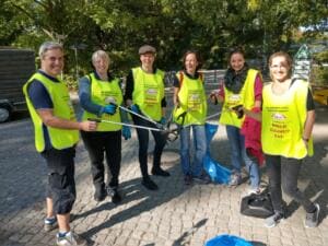 Sechs Erwachsene in gelben World-Cleanup-Day-Warnwesten stehen mit Greifzangen auf einem gepflasterten Platz unter Bäumen.
