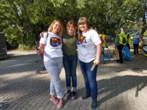 Drei lachende Frauen, zwei in World-Cleanup-Day-Shirts, stehen Arm in Arm auf einem Platz, dahinter Helfer in Warnwesten.