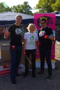 Zwei Männer in World-Cleanup-Day-Shirts und eine Frau im „Berlin Macher“-Shirt stehen mit roten Handschuhen vor einem pinken Roll-up.