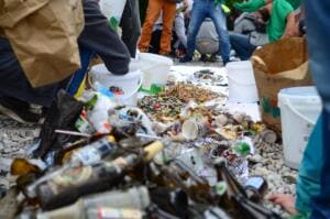 Auf einer weißen Plane am Kiesufer liegen Flaschen, Becher, Kippen und Verpackungen, ringsum sortieren Helfer in Eimer und Papiertüten.