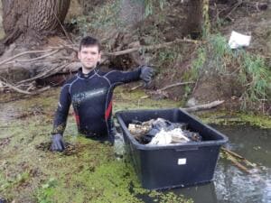 Ein Mann im Neoprenanzug steht bis zur Brust im Wasser der Itz neben einer schwarzen Kiste voller geborgenem Müll.