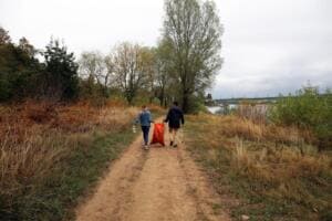 Zwei Helfer tragen einen orangefarbenen Sack und eine Greifzange einen sandigen Weg entlang durch hohes Gras zum Fluss.