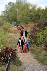 Eine lange Reihe von Helfern mit orangefarbenen Säcken und Greifzangen geht einen Weg zwischen herbstlichen Büschen entlang.