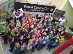 Viele Grundschulkinder in einer Schulaula winken von unten in die Kamera vor einem Banner „World Cleanup Day – 15. September 2018“.