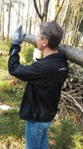 Ein Mann in schwarzer Jacke trägt einen langen Baumstamm auf der Schulter durch den Wald.