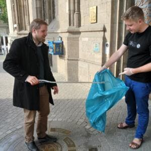 Ein Mann im Mantel hält eine lange Greifzange über einen blauen Sack, den ein zweiter Mann im schwarzen Shirt aufhält, vor einem historischen Steinportal.