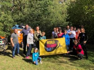 Eine Gruppe aus Erwachsenen und Kindern steht auf einer Wiese vor einem Container und hält ein gelbes World-Cleanup-Day-Banner.