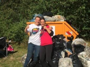 Zwei lachende Frauen, eine im World-Cleanup-Day-Shirt, zeigen den Daumen nach oben vor einem orangefarbenen Container, um den sich volle Müllsäcke stapeln.