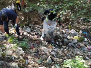 Zwei Helfer sammeln an einem Waldhang, der dicht mit Plastik, Papier und Unrat bedeckt ist, Müll in durchsichtige Säcke.