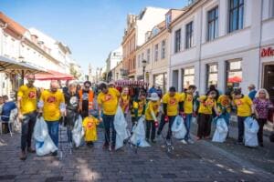 Eine Gruppe in gelben World-Cleanup-Day-Shirts zieht mit Greifzangen und weißen Säcken durch eine Fußgängerzone mit historischen Häusern.