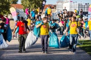Mehrere Helfer in roten und gelben Shirts tragen volle blaue und weiße Müllsäcke über einen Parkweg zur Sammelstelle.