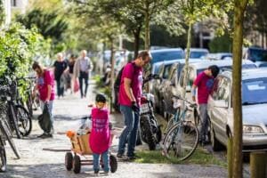 Ein kleines Kind in pinkem Shirt zieht einen Bollerwagen über einen Gehweg, daneben sammeln Erwachsene zwischen geparkten Autos und Fahrrädern Müll.