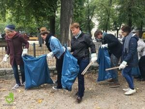 Mehrere Erwachsene in Herbstkleidung halten blaue Säcke auf und sammeln mit Greifzangen und Handschuhen Müll auf einem Kiesweg im Park.