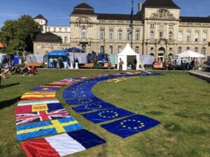 Auf einem Rasen vor einem historischen Gebäude sind im Bogen zahlreiche Flaggen europäischer Länder und der EU ausgelegt, im Hintergrund Zelte und Stände.