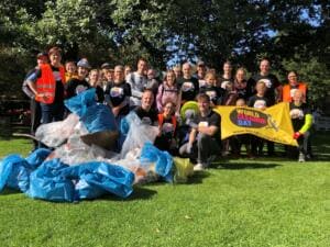 Eine große Gruppe in World-Cleanup-Day-Shirts steht und kniet auf einer Wiese hinter einem Berg voller blauer und durchsichtiger Säcke und hält ein gelbes Banner.