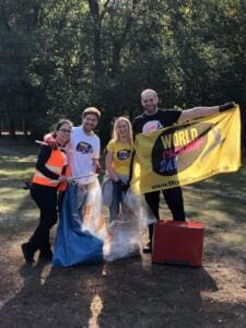 Vier Erwachsene, teils in Warnwesten und World-Cleanup-Day-Shirts, halten eine gelbe World-Cleanup-Day-Fahne in einem Park, davor ein roter Mülleimer und Säcke.