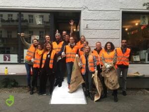 Eine Gruppe in orangefarbenen Warnwesten steht mit Jutesäcken vor einem Ladenschaufenster, einige jubeln mit Greifzangen.