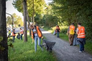 Mehrere Helfer in orangefarbenen Warnwesten sammeln mit Säcken entlang eines Rad- und Fußwegs zwischen Bäumen und einer Straße.