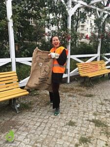 Eine lachende Frau in orangefarbener Warnweste hält einen großen Jutesack in einem Park mit weißer Pergola und gelben Bänken.