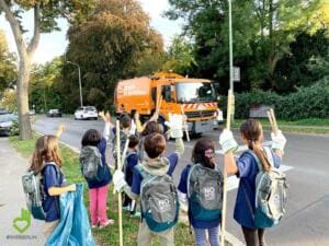 Eine Gruppe Schulkinder mit Rucksäcken und Greifzangen jubelt am Straßenrand, im Hintergrund fährt ein orangefarbenes Reinigungsfahrzeug.