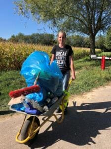 Ein Mädchen im Shirt mit der Aufschrift „Break Habits“ schiebt eine Schubkarre voller blauer Säcke, Flaschen und eines Besens über einen Weg am Maisfeld.