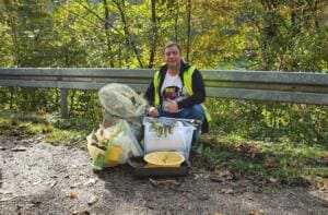 Ein Mann in Warnweste und World-Cleanup-Day-Shirt hockt an einer Leitplanke hinter Kisten, Schüsseln und Säcken mit gesammeltem Müll.
