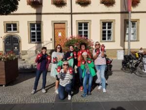 Eine Gruppe von Frauen und einem Mädchen zeigt rote Arbeitshandschuhe vor einem gelben historischen Rathaus mit Blumenschmuck.