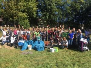 Eine sehr große Gruppe aus Erwachsenen und Kindern jubelt mit erhobenen Armen auf einer Wiese hinter vielen blauen Säcken und Eimern.