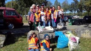 Eine Gruppe in Warnwesten steht neben einem Parkplatz hinter Kisten, Eimern und Säcken mit gesammeltem Müll, zwei Kinder sitzen vorn auf einem Stein.