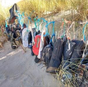 Viele angeschwemmte Schuhe, Stiefel und Flip-Flops hängen an bunten Seilresten in einer Sanddüne mit Strandhafer.