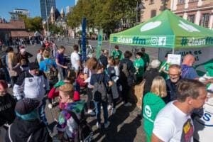 Viele Menschen drängen sich vor dem grünen #cleanffm-Stand am Mainufer, im Hintergrund Altstadthäuser und Hochhäuser.