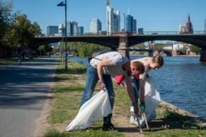 Ein Mann und eine Frau bücken sich mit Greifzangen und weißen Säcken am Mainufer, im Hintergrund Brücke und Skyline.