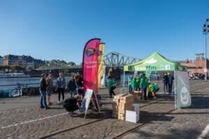 Am Mainufer stehen ein grüner #cleanffm-Pavillon, Beachflags und Kartons, im Hintergrund der Eiserne Steg und Menschen.