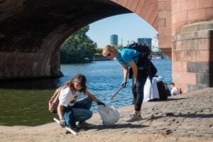 Zwei Frauen sammeln mit Greifzangen unter einem Sandsteinbogen der Alten Brücke direkt am Wasser des Mains.