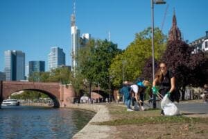Mehrere Helfer sammeln auf einem Grünstreifen am Main, im Hintergrund die Frankfurter Hochhäuser und der Dom.