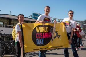 Drei Erwachsene in World-Cleanup-Day-Shirts halten eine große gelbe World-Cleanup-Day-Fahne vor einem Ausflugsschiff.
